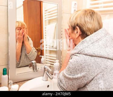 Senior woman in bathrobe touching her soft face skin, looking in mirror at home Stock Photo