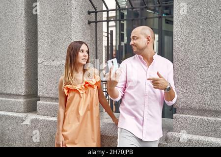 Content stylish boyfriend sharing mobile phone with girlfriend while standing near building in city in summer Stock Photo