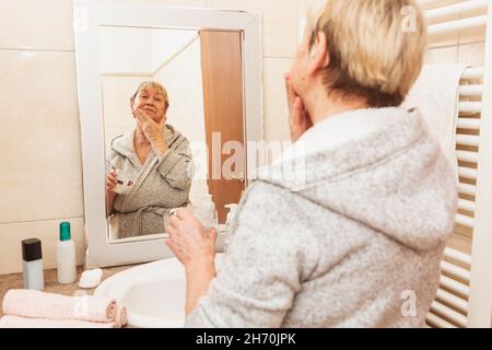 Senior woman in bathrobe touching her soft face skin, looking in mirror at home Stock Photo