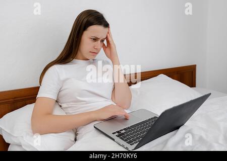 Young serious woman working at home sit in bed with laptop, nervous breakdown, waiting for interview, exam, medical test result Stock Photo