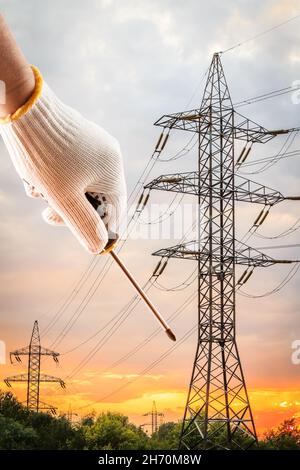 Hand dressed in a white glove holding a screwdriver with high voltage power transmission towers at sunset on the background. Stock Photo