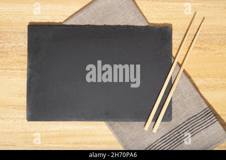 Flat lay of Blank Black slate Tray, chopsticks and linen napkin on wooden table. Template Mock up Stock Photo