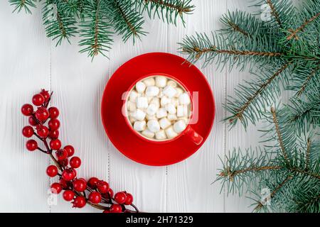 Red cup of coffee with marshmallow and Christmas tree on white wooden table. New Year decorations, spruce branches and red holly berries. Winter seaso Stock Photo