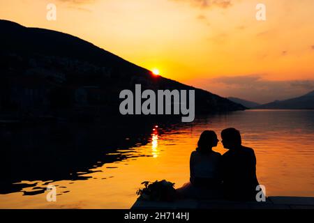 Silhouettes of couple sit on the pier above the water against the backdrop of mountains and sunset Stock Photo