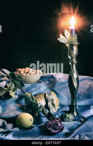 Still life with lighting candle, dry rose, lemon, old pastry and hazelnuts in a bowl on blue and dark background Stock Photo