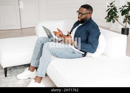 Focused African-American guy using laptop for video call sitting on the sofa at home, full length. Black freelancer man in casual shirt and glasses taking online, tutor giving online language classes Stock Photo