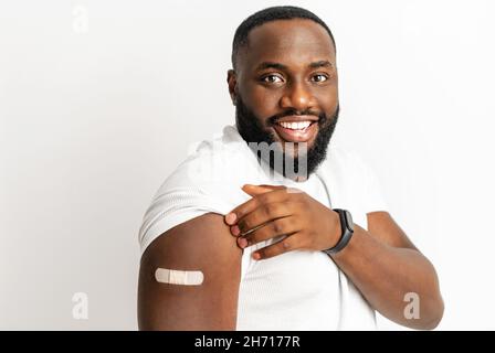 Cheerful vaccinated African-American man showing arm with medical patch and laughs, black male getting vaccine dose against covid, plaster on her shoulder, isolated on white. Healthcare concept Stock Photo