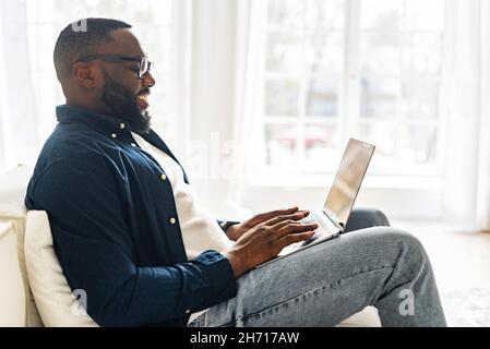 Side view at young modern African-American freelancer guy using laptop, black man in casual shirt and glasses typing sitting on the sofa, working remotely from home, web surfing, sending emails Stock Photo
