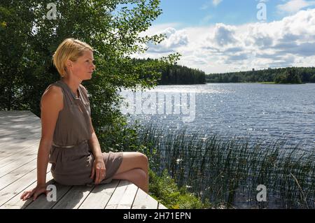 A young woman sitting on a wooden deck looking at a lake Stock Photo