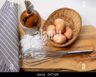 A kitchen whisk, flour, eggs and a bird feather in a ceramic container on a wooden cutting board Stock Photo