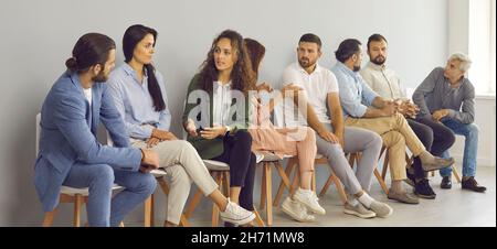 Group of diverse people communicate with each other sitting on chairs in line in the waiting room. Stock Photo
