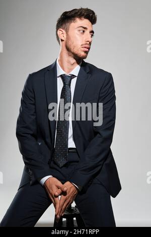Young man portrait seated in a stool wearing a dark blue suit looking to his left. Stock Photo