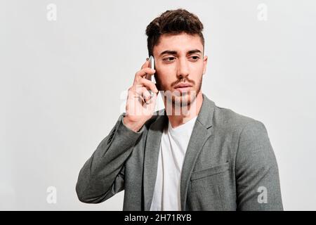 Serious young man wearing jeans, a white tshirt and a grey blazer walking in front of a white wall while talking with someone on his mobile phone Stock Photo