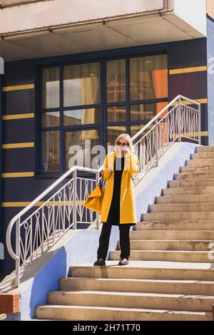 Elegant modern Woman with blond hair in a bright yellow coat and sunglasses walks along a city street. Urban style of modern woman walking shopping Stock Photo