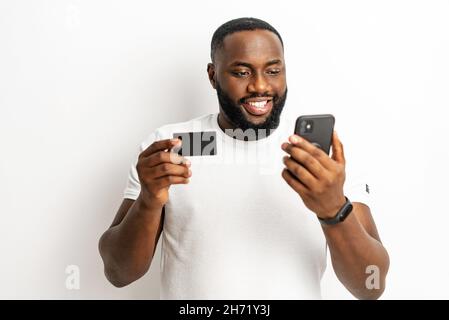 E-banking, money transferring online. Cheerful African-American man holding credit card and smartphone in his hands isolated on white, black guy using mobile app for online payment, shopping Stock Photo