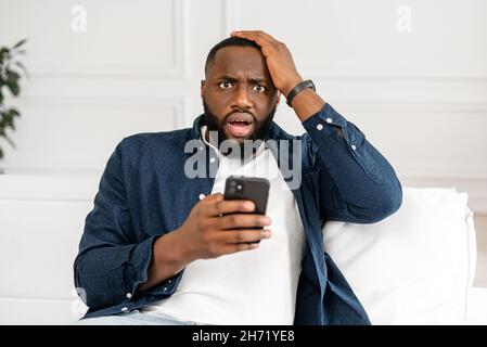 African-American guy amazed with bad news received on the smartphone, holds a phone and staring at the screen with mouth opened, holds his head and feels shock, worrying about what saw on the net Stock Photo