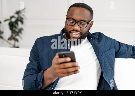 Close-up portrait of millennial bearded African-American man in glasses using smartphone for spending leisure time, dating, chatting in social networks, black cheerful guy watching at phone screen Stock Photo