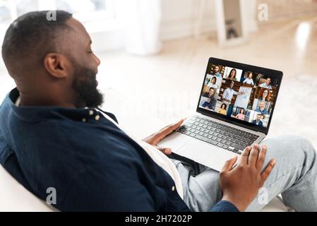 Back view on the laptop screen with many profiles on it, an African-American guy is using computer app for video meeting, online conference with coworkers, colleagues, virtual briefing Stock Photo