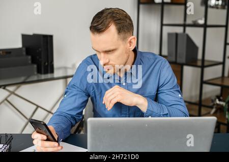 Concentrated serious man received notification on his smartphone, reading message sitting at the desk in front of the laptop, male freelancer using phone working on the distance Stock Photo