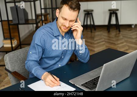 Smiling man talking phone and work from home with laptop computer, freelance male writing on notebook and speak on smartphone, guy takes notes during phone conversation Stock Photo