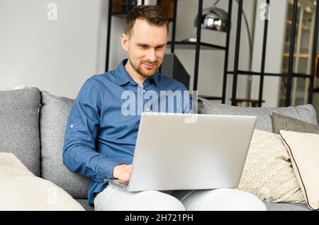 Smiling young bearded man is using a laptop at home, a guy looks at the device screen with interest sitting on the couch, websurfing, watching news feed, works remotely from home Stock Photo