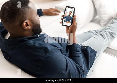 Two diverse male friends or colleagues talking on the distance. African-American man lying on white sofa in modern apartment, using smartphone for video call, talking with indian guy Stock Photo