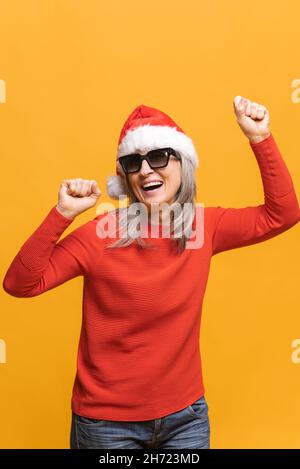 Overjoyed middle-aged gray-haired woman in red santa hat and sunglasses dancing, makes movements to music, smiles positively, being in high spirit. Carefree mature lady dances isolated on yellow Stock Photo