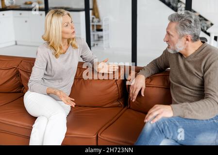 Irritated middle-age couple has hard debate at home, sitting on the sofa and arguing, annoyed woman talking to angry man, mature spouses has problems in marriage Stock Photo