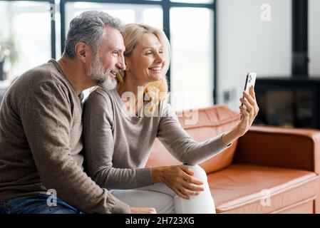 Smiling middle-aged couple using smartphone sitting on the comfortable sofa at home, cheerful spouses using mobile phone, watching movies, websurfing, making video call to grown kids Stock Photo