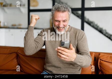 Senior grey-haired man is using smartphone for watching sports soccer match or play video game sitting on comfortable sofa at home. Cheerful mature male raising fist up in victory gesture, triumph Stock Photo