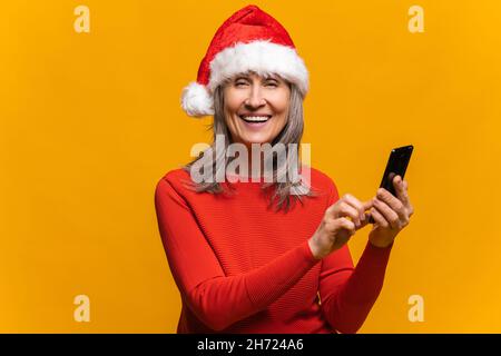 Close-up portrait of hilarious senior woman in santa hat using smartphone isolated on yellow, middle-aged gray-haired lady sending New Year congratulations, texting online