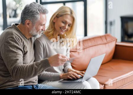 Cheerful middle-aged couple using laptop for making purchases in online shopping, mature spouses making order and using debit card for paying, making online transaction, transfer money to grown child Stock Photo