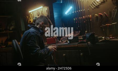 Young blacksmith man with workwear standing up and get proper tool for strip the cable, sitting down and working on workbench in his workshop Stock Photo