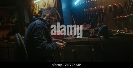Young blacksmith man with workwear standing up and get proper tool for strip the cable, sitting down and working on workbench in his workshop Stock Photo