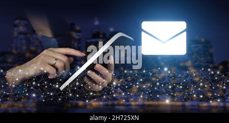 Email marketing concept. Sending newsletter. Hand touch white tablet with digital hologram Email and sms sign on city dark blurred background. Sending Stock Photo