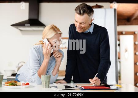 Smiling mid aged couple using cellphone and making notes in planner in kitchen Stock Photo