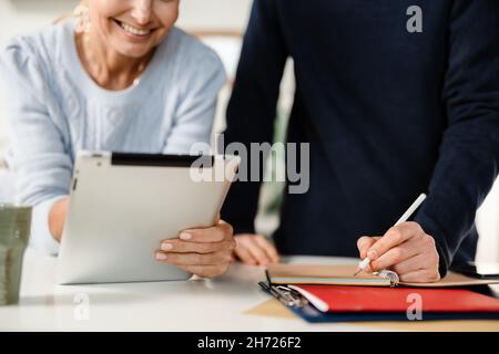 Close up of a smiling mid aged couple using digital tablet and making notes in planner in kitchen Stock Photo