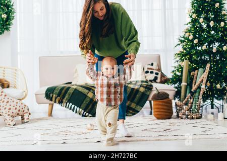 The child with the support of his mother takes the first steps. Christmas happy family moments. Stock Photo