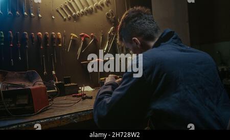 Young workshop owner man with workwear, sitting on chair and solder on workbench Stock Photo
