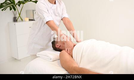 Unrecognizable masseuse giving shoulder massage to a middle aged man relaxing on massage table in luxury white spa beauty clinic. Therapeutic body mas Stock Photo