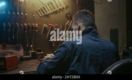 Young workshop owner man with workwear, sitting on chair and solder on workbench Stock Photo