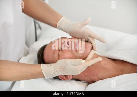 Close-up of handsome mature 50 years old man relaxing on massage table while getting professional facial lifting massage at spa. Anti-aging, rejuvenat Stock Photo