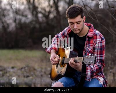 Handsome young man plays acoustic guitar on the natural park. Stock Photo