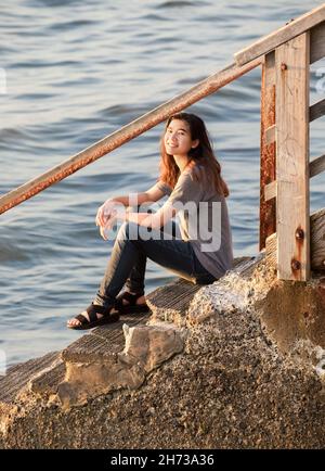 Biracial Asian teen girl relaxing on stone steps leading down to water Stock Photo