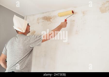 A man in a paper cap makes repairs at home, prime the walls with a paint roller. Renovation diy new home painting wall. Stock Photo