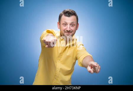 Playful smiling young man touching you over blue background, dresses in yellow shirt. Amusing guy. Studio shot Stock Photo