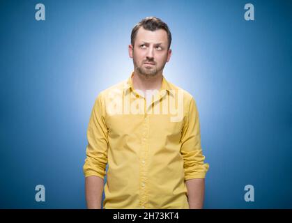 Consider man thinking while looking up to blank space over blue background, dresses in yellow shirt. Half length of pensive guy. Studio shot Stock Photo