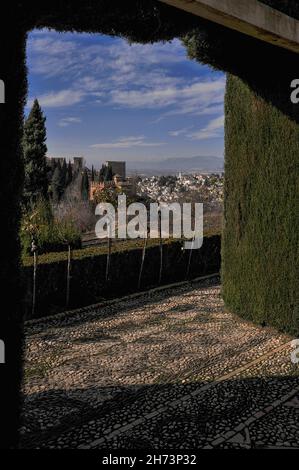 The Alhambra royal palace and Moorish Albaicín District of the city of Granada, Andalusia, Spain, seen through a topiary arch above a river pebble mosaic pavement in the gardens of the Generalife former sultans’ summer palace Stock Photo