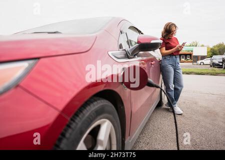 Woman with smartphone next to charging red electric car Stock Photo