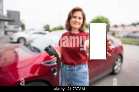 A woman on the background of a red electric car shows a blank smartphone screen to the camera. You can add your content here Stock Photo